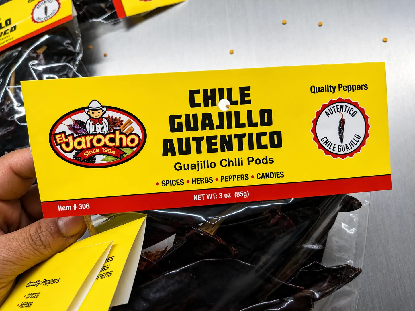 Close-up of a hand holding a yellow El Jarocho header label reading 'Chile Guajillo Autentico' stapled to a clear bag of dried chili pods.