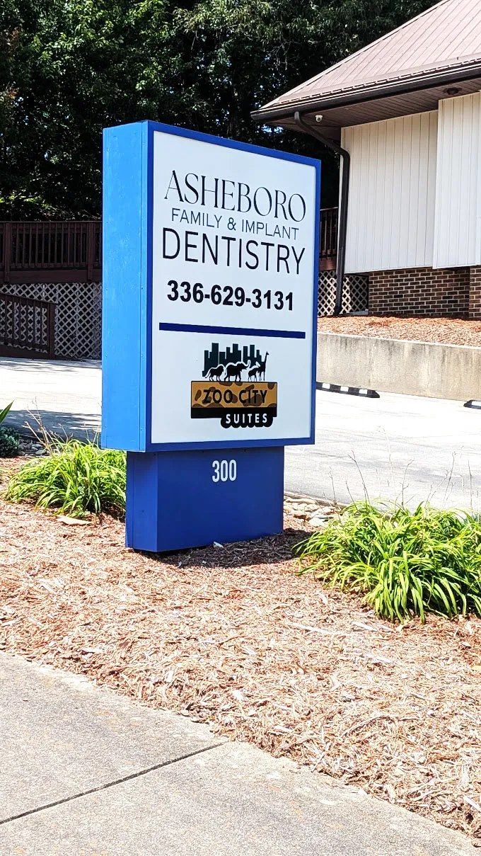 Blue and white pylon sign for Asheboro Family and Implant Dentistry standing in landscaped mulch beside a driveway.