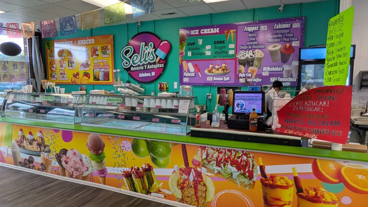 Interior counter view of Beli's Nevería y Antojitos showing full-color counter wrap with ice cream photography, illuminated menu boards for ice cream, frappes, and paletas, and wall graphics on a teal wall.