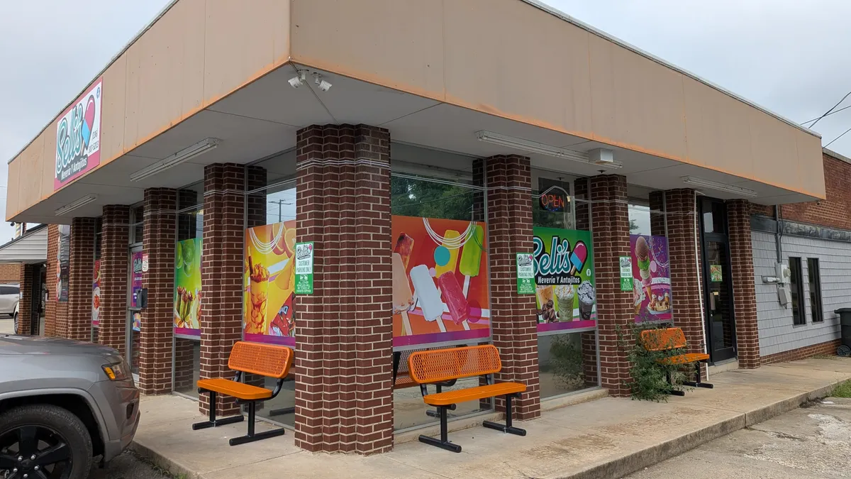 Corner view of Beli's Nevería y Antojitos storefront covered in full-color window graphics showing paletas, fruit drinks, and ice cream against orange and pink backgrounds, with orange benches outside.