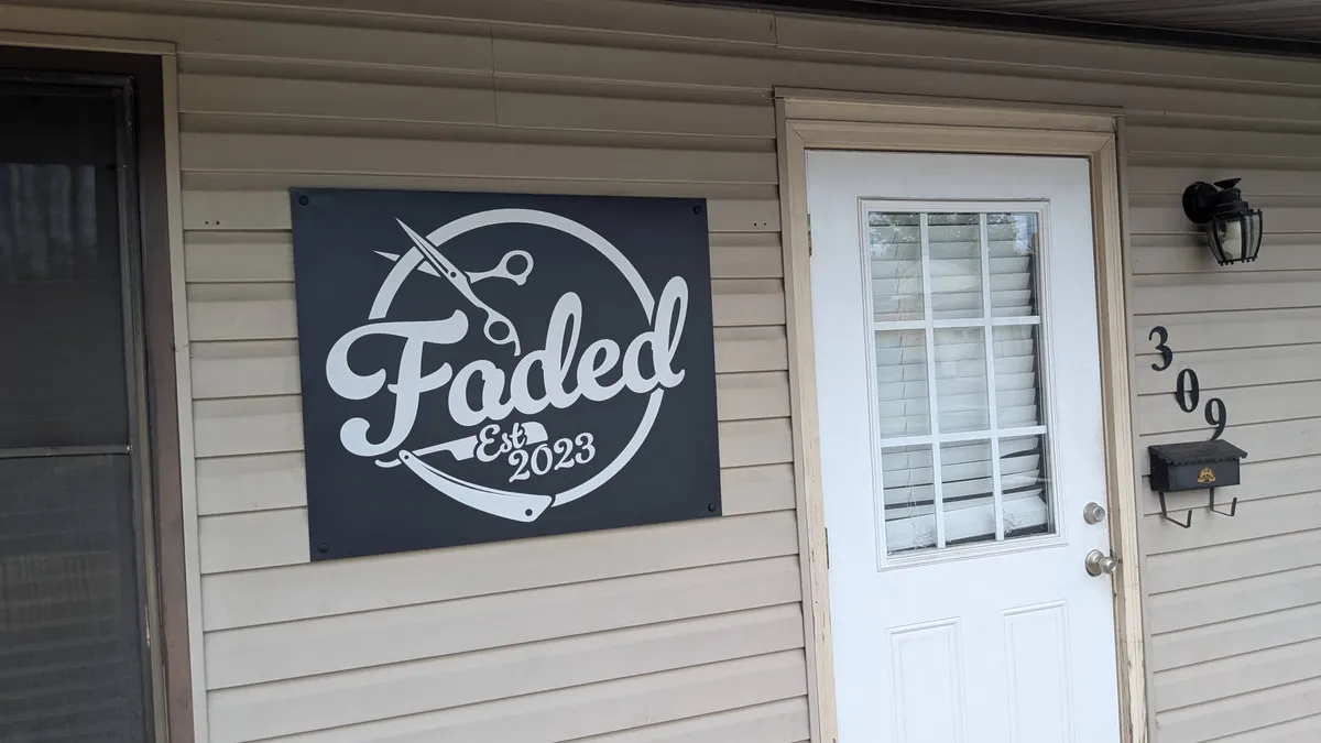 Dark charcoal flat panel sign for Faded Est. 2023 barbershop with white script lettering, scissors, and straight razor graphic, mounted on tan vinyl siding.