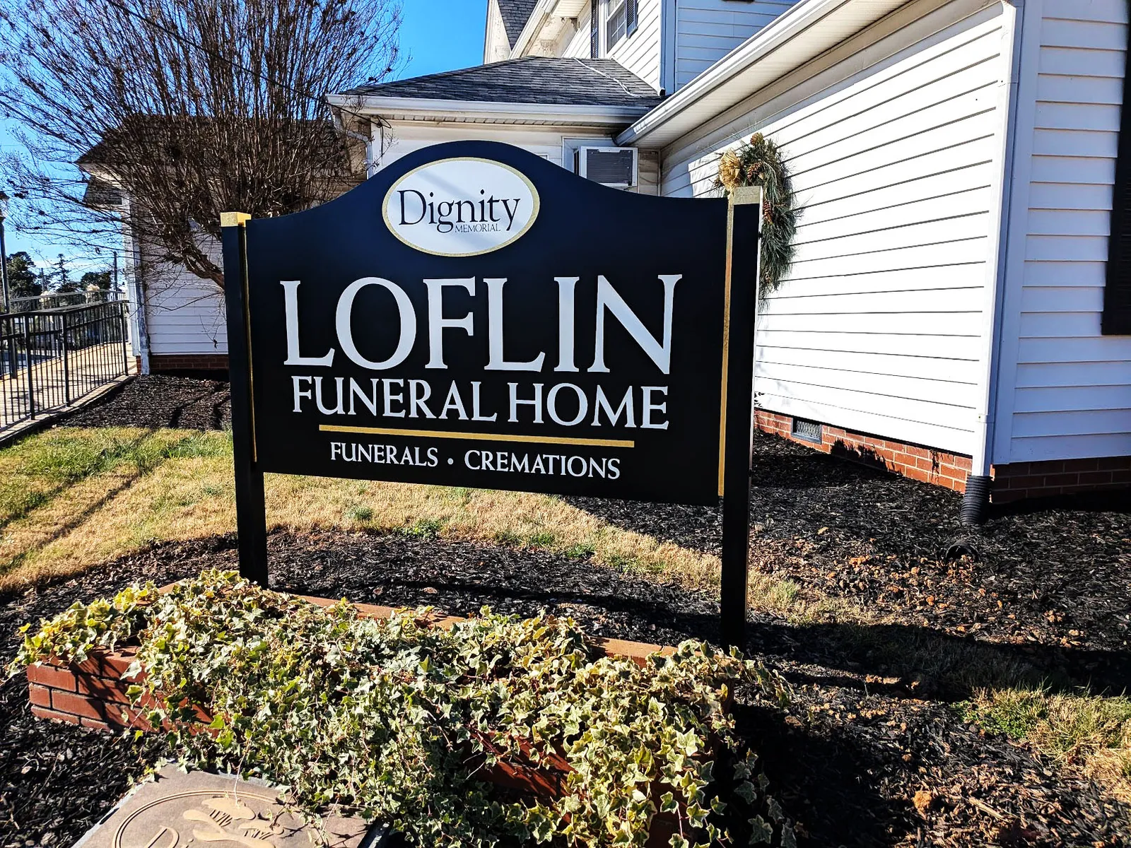 Black and gold post-and-panel sign for Loflin Funeral Home in front of a white building on a sunny day.