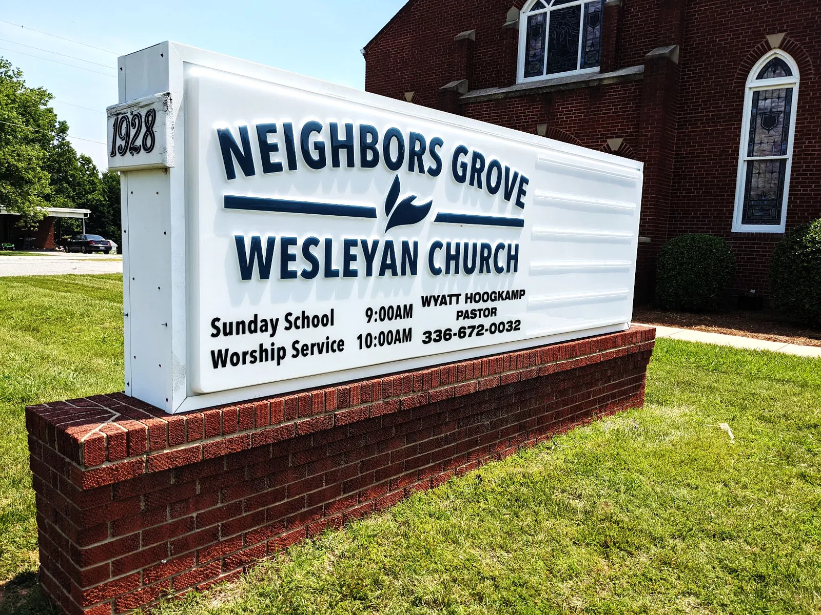 White monument sign for Neighbors Grove Wesleyan Church with service times, mounted on a red brick base in front of the church.