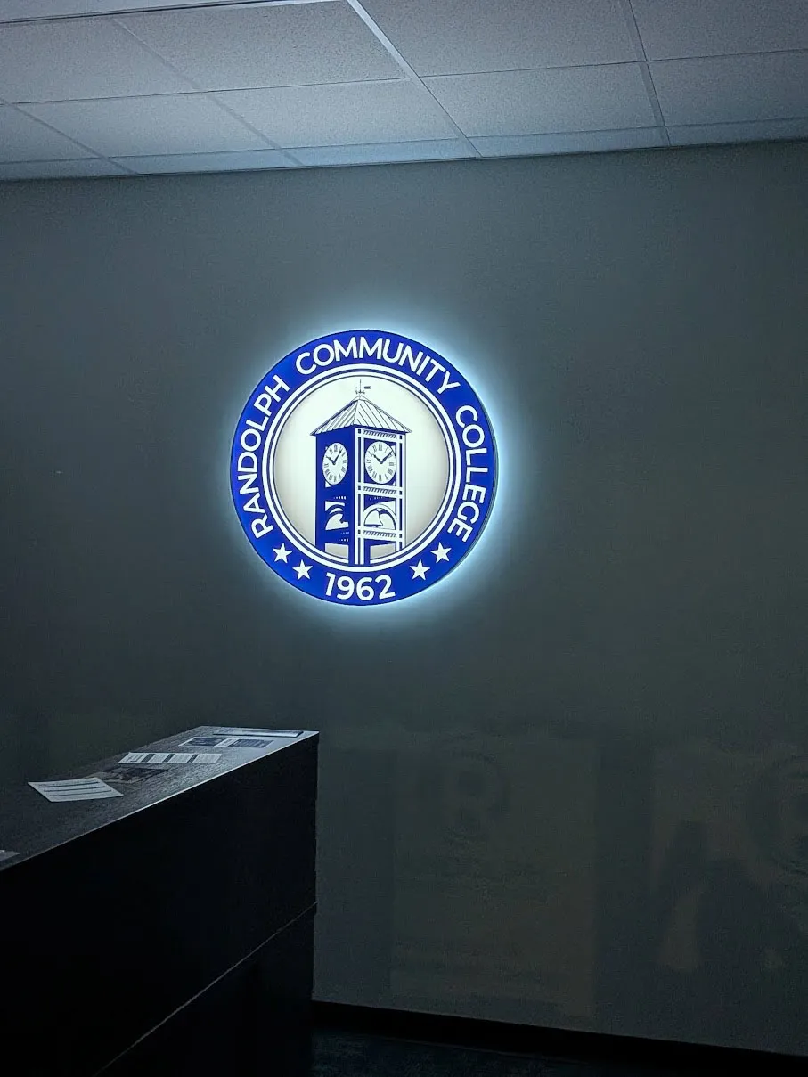 Blue and white backlit circular wall sign for Randolph Community College Est. 1962, showing a clock tower inside a ring of text and three stars, glowing with a white halo on a dark interior wall.