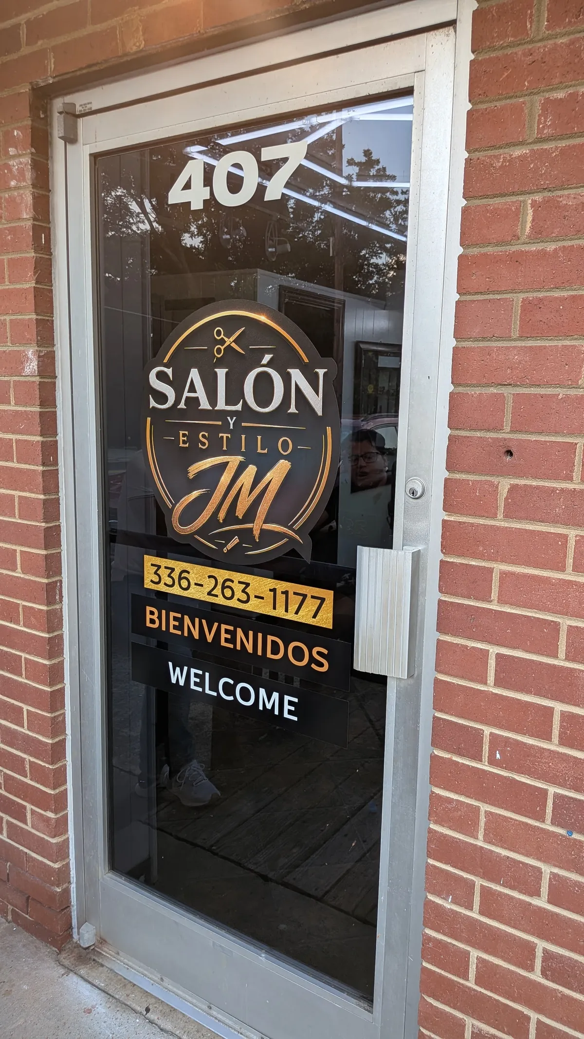 Gold and black circular Salón y Estilo JM logo with scissors graphic applied to a glass door, phone number and Bienvenidos / Welcome text below, in Asheboro, NC.