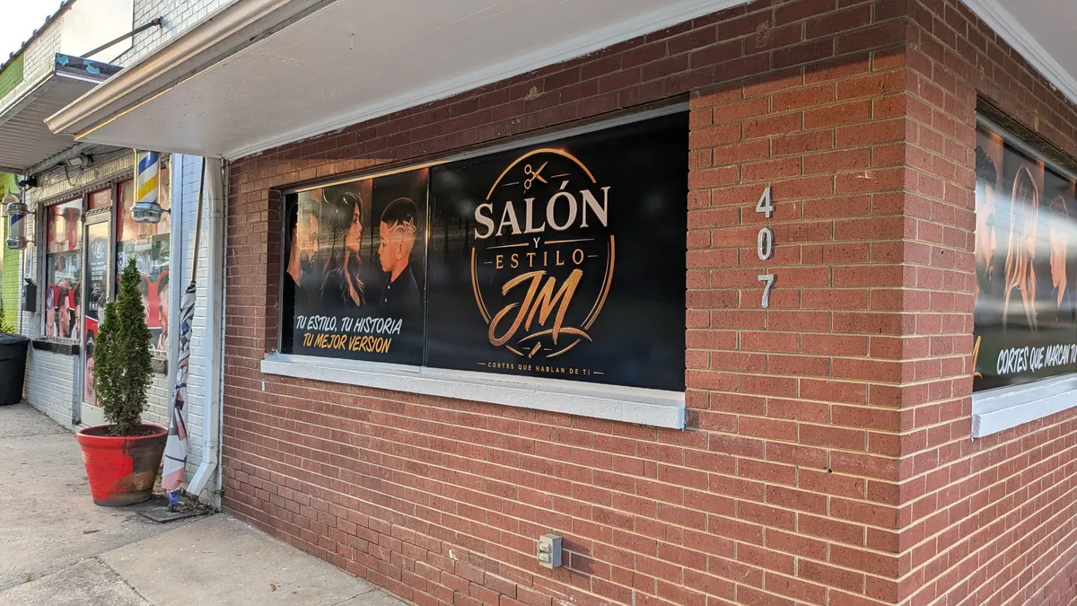Full exterior view of Salón y Estilo JM storefront with large black window graphics showing the salon logo, gold script, and model photography across two windows on a brick building in Asheboro, NC.