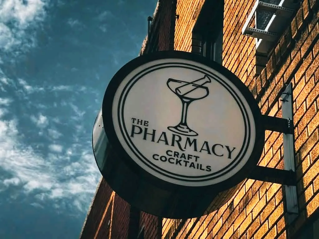 Round black-and-white blade sign for The Pharmacy Craft Cocktails mounted on a brick building against a dramatic cloudy sky.