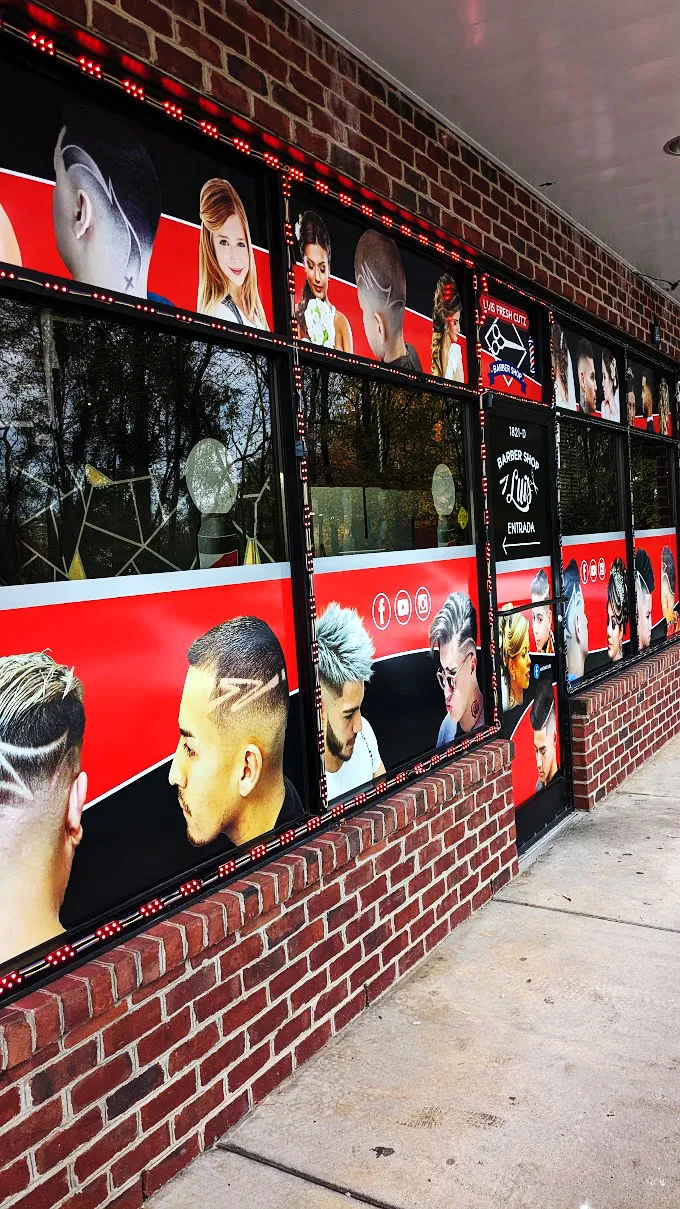 Exterior of a brick storefront with multiple windows fully wrapped in red and black photo-collage graphics for Luis Fresh Cutz Barber Shop, showing haircut photos and salon branding with red LED string lights framing the panes.