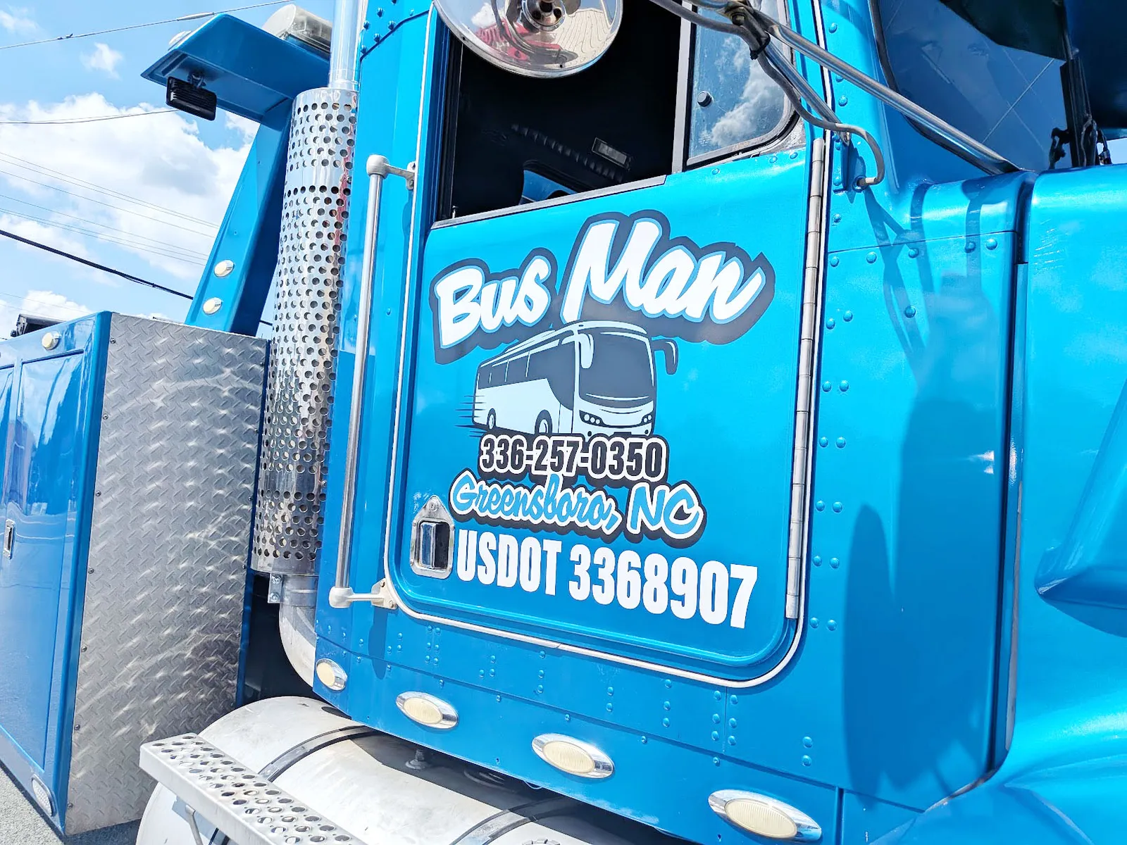 Close view of a bright blue semi truck door with white Bus Man script lettering, a bus illustration, and Greensboro NC details.