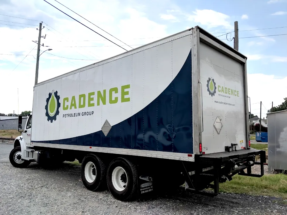 Three-quarter rear view of a white box truck with a bold navy wave stripe and large Cadence Petroleum Group logo in lime green and gray covering the full side and rear door.