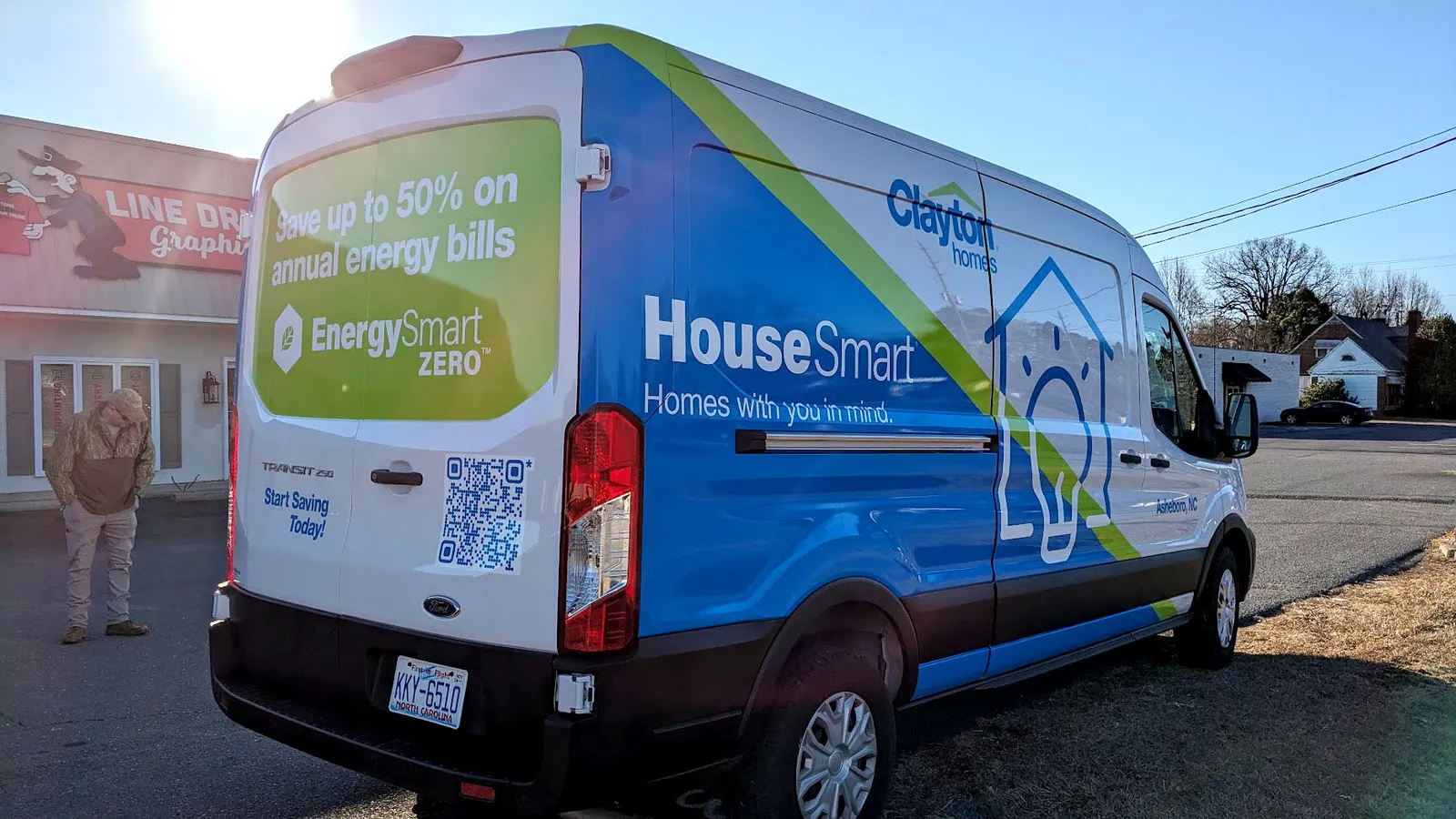 Rear three-quarter view of a Ford Transit van in a full blue, green, and white wrap promoting Clayton Homes' HouseSmart energy program.