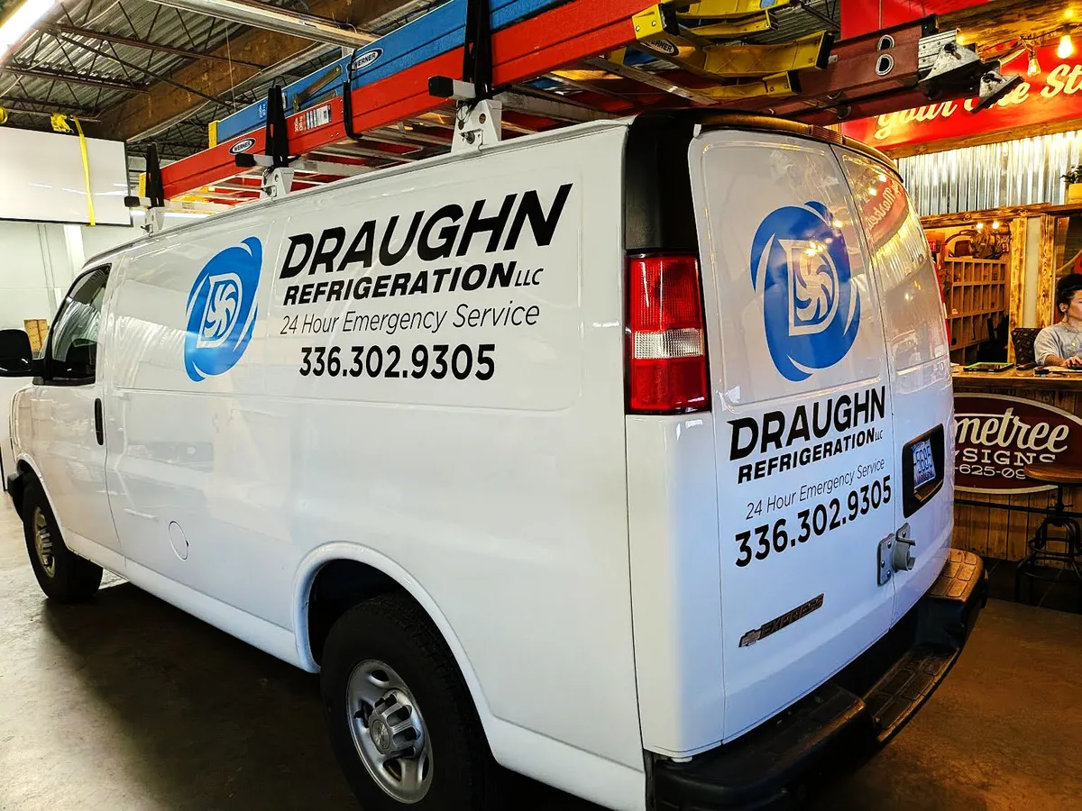 Three-quarter rear view of a white Chevy Express van lettered for Draughn Refrigeration LLC with a blue swirl logo, 24 Hour Emergency Service, and phone number on both the side and rear door, photographed inside a sign shop.