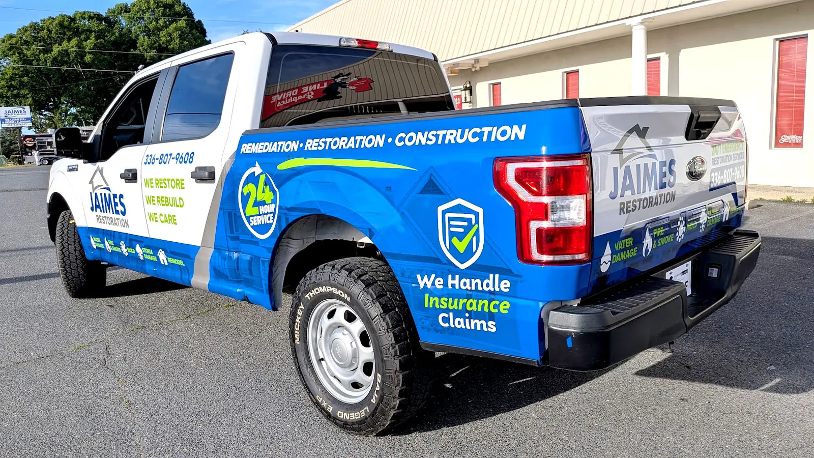 Rear three-quarter view of a pickup truck wrapped in blue and white Jaime's Restoration graphics with a 24-hour service badge.