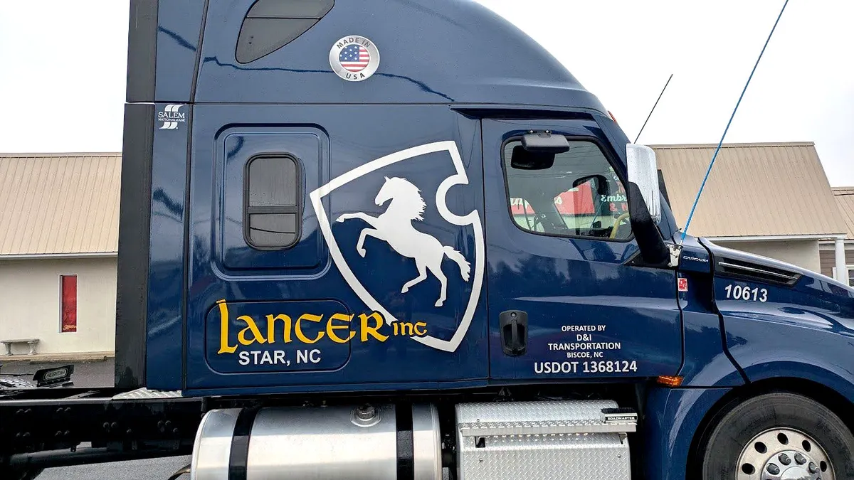 Side view of a navy blue Freightliner Cascadia sleeper cab with a large white rearing-horse shield and gold script Lancer Inc lettering, located in Star, NC.