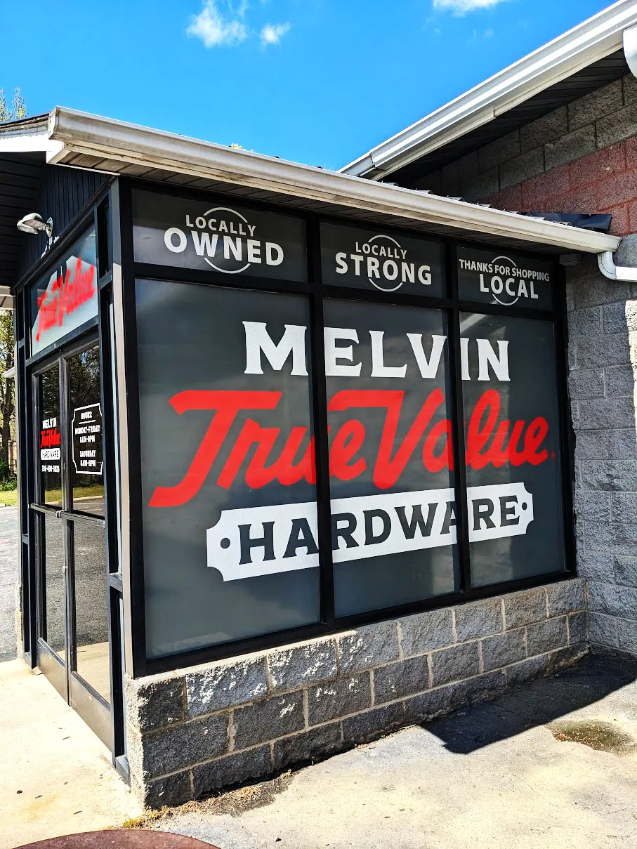 Storefront windows covered in gray perforated graphics with red and white Melvin True Value Hardware lettering and locally-owned slogans.
