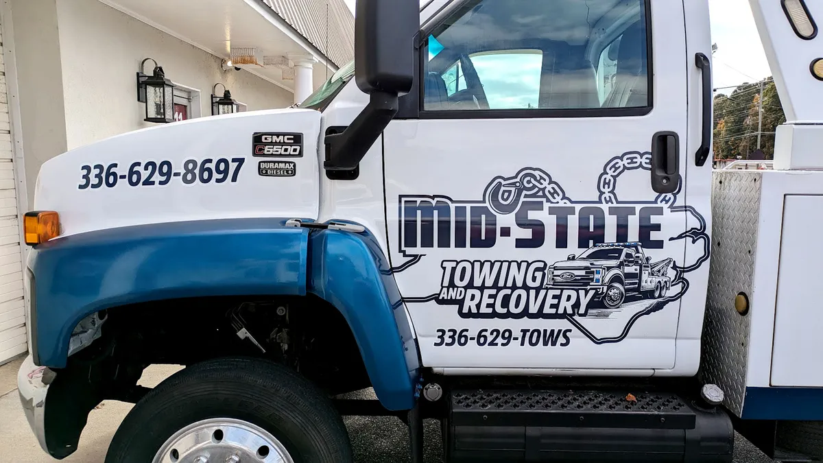 White and blue GMC C6500 cab door with Mid-State Towing and Recovery graphics: bold lettering surrounded by illustrated chain links and a wrecker truck artwork, with 336-629-TOWS phone number.
